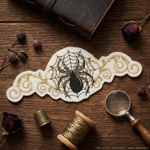May include: An embroidered patch featuring a black spider in a web, with gold filigree accents. The patch is cream-colored and has the text "THE GILDED ARACHNID - Artisan Embroidery". The patch is on a wooden surface.