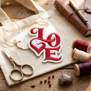 Op de afbeelding: Een witte patch met het woord "LOVE" geborduurd in rood, geplaatst op een beige draagtas. De afbeelding bevat een kleine schaar, spoelen garen, een leren notitieboek en kleine rode kralen, allemaal gerangschikt op een houten oppervlak.