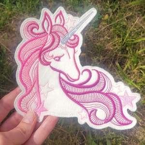 Könnte beinhalten: Gestickter Einhorn-Aufnäher mit weißer Basis und rosa Details. Das Einhorn hat ein silbernes Horn, eine rosa Mähne und einen rosa Schweif mit Sternakzenten. Der Aufnäher ist detailliert gestickt.