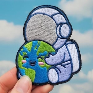 Könnte beinhalten: Gestickter Aufnäher mit einem Astronauten in einem blauen Raumanzug, der eine lächelnde Erde umarmt. Der Astronaut hat einen grauen Helm. Die Erde ist blau und grün mit einem lächelnden Gesicht und rosa Wangen. Der Aufnäher hat einen schwarzen Rand.