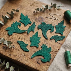 May include: A collection of dark green embroidered leaf appliques arranged on a wooden cutting board. The leaves vary in size and design, with detailed stitching. Green thread spools and needles are also present, suggesting a crafting theme.