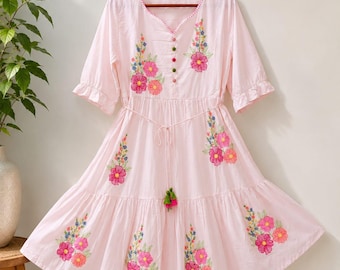 May include: A light pink knee-length dress with half-length ruffled sleeves and a V-neckline. The garment features colorful floral embroidery, a central row of small decorative buttons, and a thin waist tie with green and pink tassel accents.