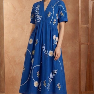 May include: A vibrant blue midi dress featuring a V-neckline, short puff sleeves, and an empire waist. The fabric is adorned with white and muted-tone embroidered floral and botanical patterns, including delicate leaf sprigs and circular blooms.
