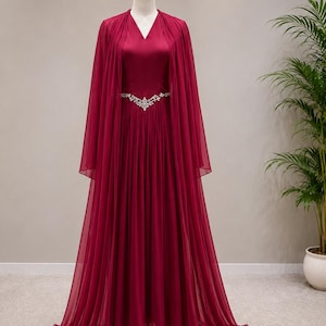 May include: A floor-length, deep burgundy gown featuring a V-neckline and a flowing, sheer cape overlay. The dress is cinched at the waist with a decorative silver-toned belt adorned with clear crystals, and the skirt has soft, vertical pleats.