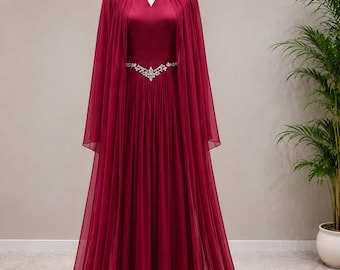 May include: A floor-length, deep burgundy gown featuring a V-neckline and a flowing, sheer cape overlay. The dress is cinched at the waist with a decorative silver-toned belt adorned with clear crystals, and the skirt has soft, vertical pleats.