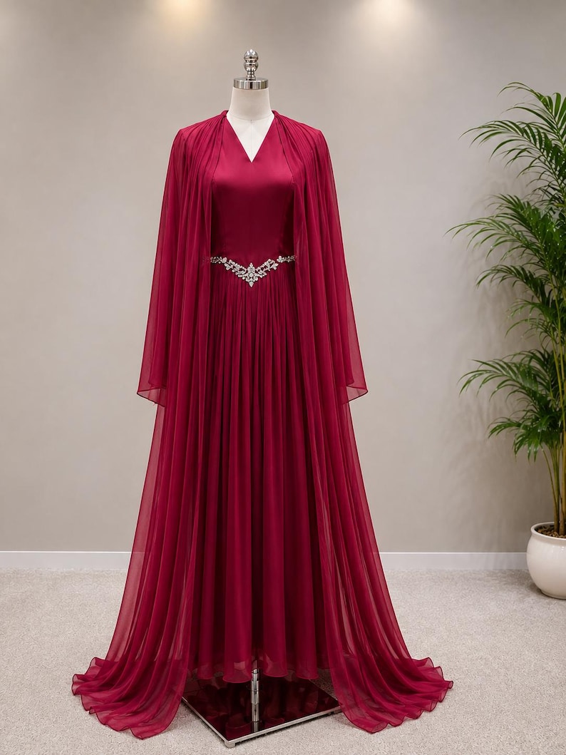 May include: A floor-length, deep burgundy gown featuring a V-neckline and a flowing, sheer cape overlay. The dress is cinched at the waist with a decorative silver-toned belt adorned with clear crystals, and the skirt has soft, vertical pleats.