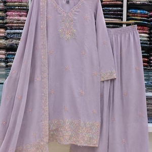 May include: A light purple three-piece outfit consisting of a long-sleeved tunic, wide-leg trousers, and a matching dupatta. The tunic features intricate floral embroidery around the neckline, cuffs, and hem, with scattered floral motifs throughout.