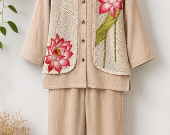 May include: A beige two-piece set consisting of a long-sleeved button-down shirt and matching trousers. The shirt features a cream-colored crochet overlay on the front panels, decorated with embroidered pink lotus flowers and green stems.