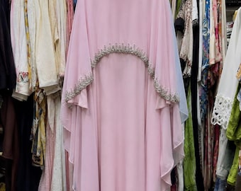 May include: A floor-length light pink dress featuring a high neckline and a layered cape-style overlay. The collar and the curved hem of the cape are embellished with intricate silver beadwork and clear crystals, creating a decorative border.
