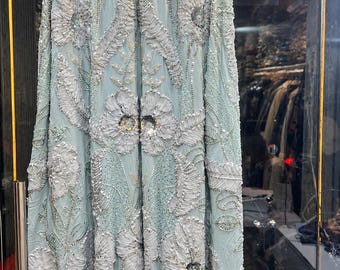 May include: A floor-length light blue gown featuring an intricate high neckline adorned with silver beads. The fabric is heavily embellished with raised white floral embroidery, silver threadwork, and scattered clear sequins throughout the entire garment.