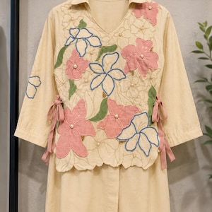 Puede incluir: Una túnica de color beige con mangas tres cuartos y cuello en V, con grandes aplicaciones de tela floral en rosa y azul, detalles de hojas verdes y pequeñas perlas blancas. Los laterales incluyen lazos de cinta rosa en la cintura.
