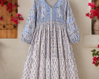 May include: A long-sleeved midi dress featuring a V-neckline and a tiered skirt. The garment is made of a light-colored fabric adorned with a repeating blue floral block print pattern, with more intricate blue floral detailing around the bodice.