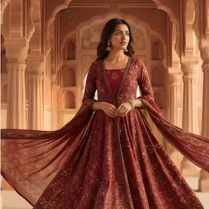 May include: A deep red floor-length dress featuring a complex gold floral pattern, a square neckline, and long sleeves. The garment includes a matching sheer shawl draped over the arms and a coordinating pair of trousers visible at the hemline.