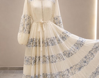 May include: A cream-colored floor-length dress featuring long bishop sleeves, a high collar, and a vertical row of small pearl-like buttons. The garment is accented with horizontal bands of blue and white floral patterns and a matching waist tie.