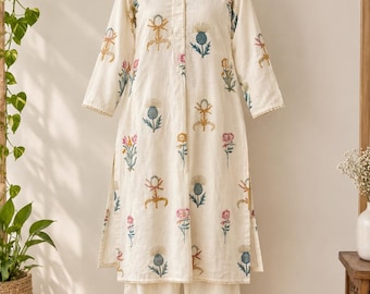 May include: A cream-colored long-sleeved tunic and matching trousers set featuring a repeating pattern of embroidered flowers in shades of blue, pink, yellow, and green. The tunic has a collared neckline, a button-down front, and side slits.