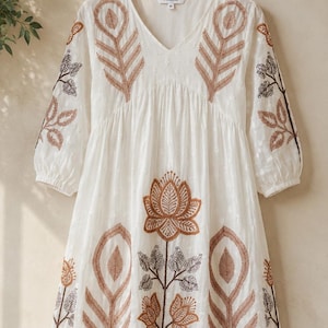 May include: A white V-neck dress featuring brown and dark grey embroidered botanical patterns, including large leaf motifs and a central lotus flower design. The dress has three-quarter length sleeves and a label reading FIXELIA size M.