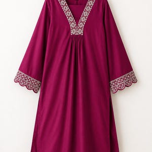 May include: A magenta tunic and matching trousers set. The tunic features long sleeves, a V-neckline, and side slits. The neckline and sleeve cuffs are decorated with white floral embroidery and a scalloped edge. The set is displayed on a hanger.