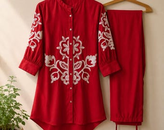 May include: A bright red tunic shirt with a mandarin collar, button-down front, and three-quarter length sleeves, featuring white floral embroidery on the chest and sleeves, paired with matching red trousers hanging on a wooden hanger.