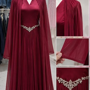 May include: A floor-length burgundy gown featuring a V-neckline, a pleated skirt, and a sheer cape overlay. The waist is accented with a silver-toned, V-shaped embellishment adorned with clear crystals. The fabric has a smooth, satin-like finish.