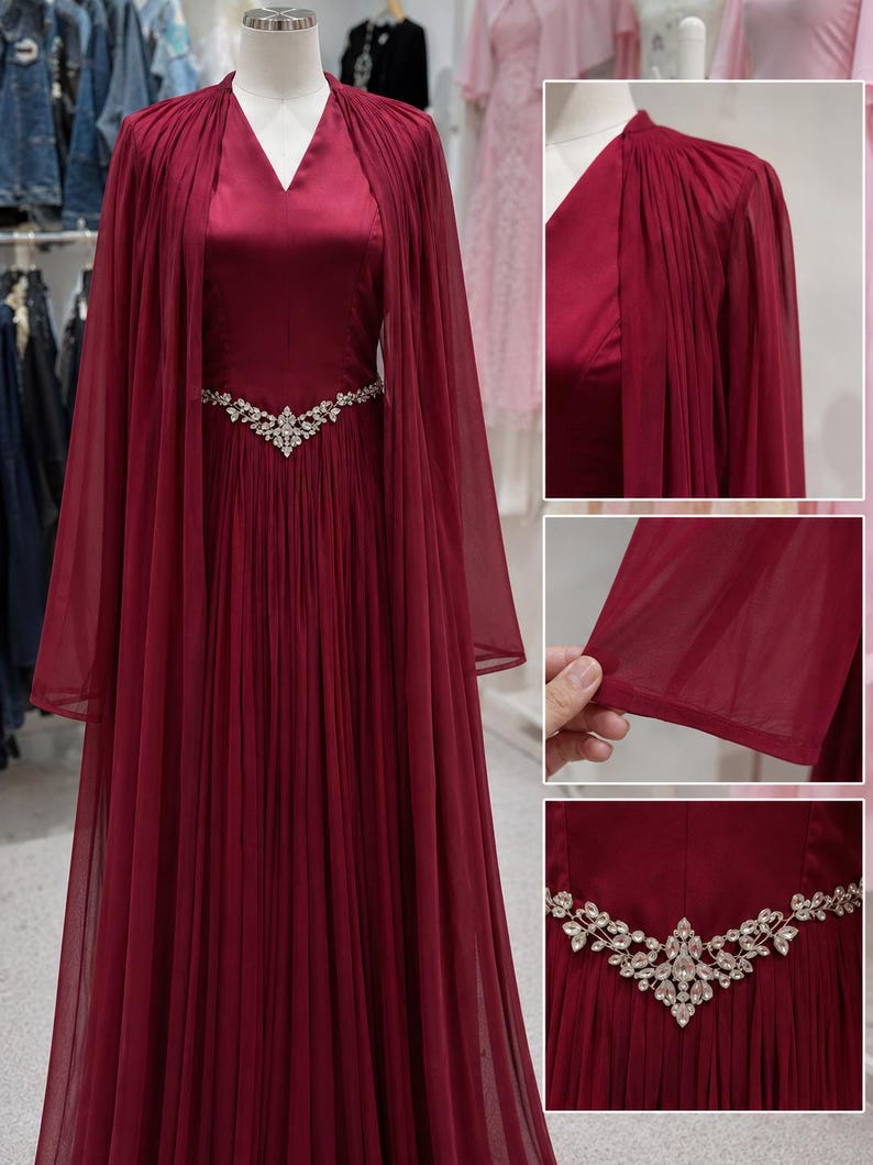 May include: A floor-length burgundy gown featuring a V-neckline, a pleated skirt, and a sheer cape overlay. The waist is accented with a silver-toned, V-shaped embellishment adorned with clear crystals. The fabric has a smooth, satin-like finish.