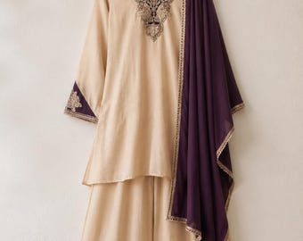 May include: A beige tunic and wide-leg pants set featuring a deep purple scarf draped over one shoulder. The tunic has a V-neckline with intricate dark purple and gold embroidery, and the sleeve cuffs are accented with matching purple fabric.