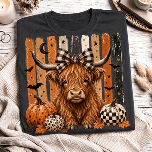 Op de afbeelding: Donkergrijze t-shirt met een illustratie van een Schotse hooglander met een buffelruit strik, hoorns en pompoenen. Het ontwerp bevat oranje en zwarte strepen, vleermuizen, sterren en een maan, wat een Halloween-thema oproept.