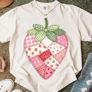 以下が含まれることがあります： パッチワークのイチゴのデザインが施された白いTシャツ。イチゴは、ピンク、赤、緑の色合いのギンガム、水玉、花柄など、さまざまな生地パターンで構成されています。シャツはクルーネックで半袖です。