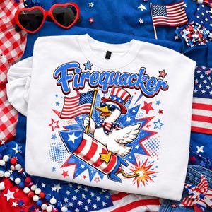 May include: White sweatshirt with a graphic of a duck wearing sunglasses, a patriotic hat, and bow tie, riding a rocket. The text "Firequacker" is above the duck. The design includes American flags and stars in red, white, and blue.