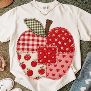 May include: White t-shirt featuring a large, patchwork apple design. The apple is composed of various red fabrics with patterns like gingham, floral, and hearts. A green gingham leaf and brown stem complete the design. The shirt has a V-neck.