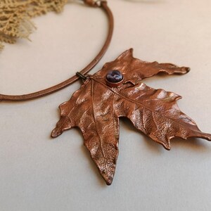 May include: A copper-coloured leaf pendant necklace. The pendant is a detailed maple leaf shape with a small, round, purple gemstone at its centre. The necklace has a black cord and a copper-toned metal chain.