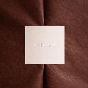 May include: A square, off-white card with the embossed text "æsthe nori" centered within a thin, debossed border. The card rests on a dark brown, textured surface with visible folds and creases.