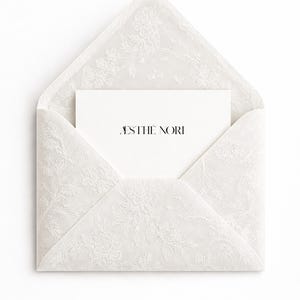 Puede incluir: Un sobre blanco con un delicado diseño de encaje floral, abierto para revelar una tarjeta blanca. La tarjeta presenta el texto "ÆSTHE NORI" en una fuente minimalista. El diseño es limpio y elegante, adecuado para papelería.