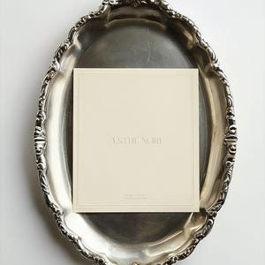 May include: A silver-toned, ornate oval tray holds a cream-colored card with the text "ASTHE NORI" and "MADE IN ITALY" in a serif font. The tray has a decorative, scalloped edge and sits on a white surface.