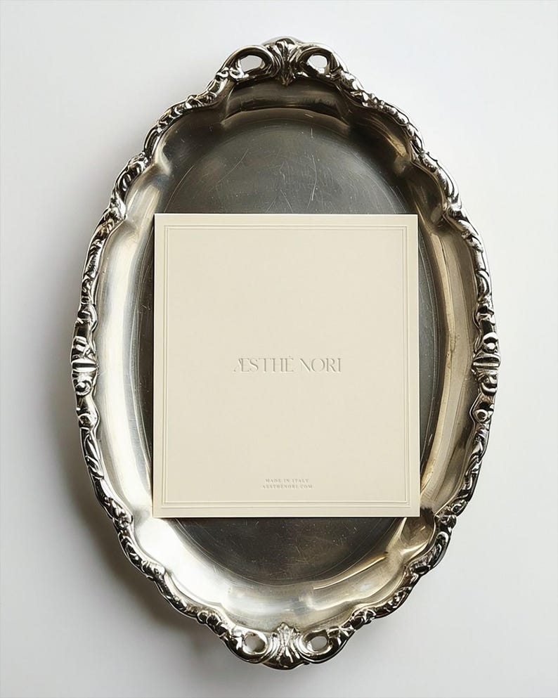 May include: A silver-toned, ornate oval tray with a cream-colored card featuring the text "ESTHE NORI." The card is centered on the tray, which has a decorative, scalloped edge. The card also has the text "MADE IN ITALY" and a website address.