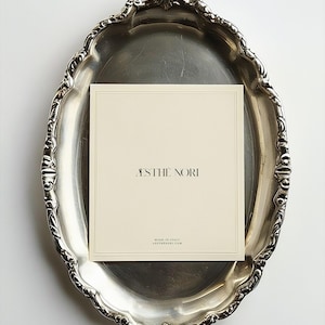 May include: A silver-colored, ornate, oval tray holds a cream-colored card with the text "ASTHE NORI" in black. The card also includes the text "MADE IN ITALY" and a website address.