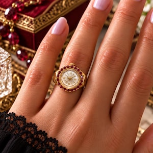 May include: A gold ring featuring a round watch face as the centerpiece, surrounded by a circular border of small, vibrant red gemstones. The watch dial is white with gold-toned numerals and hands, and a small winding crown at the top.