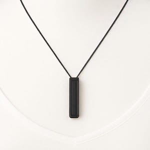 Puede incluir: Un collar negro minimalista con un colgante rectangular. El colgante cuelga de un fino cordón negro. El colgante es negro mate liso, y el cordón es de un material fino y tejido. El collar se muestra sobre un fondo blanco.
