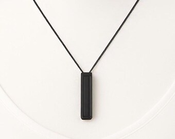 May include: A minimalist black necklace featuring a rectangular pendant suspended from a thin black cord. The pendant is a solid, matte black, and the cord appears to be a fine, woven material. The necklace is displayed against a white background.