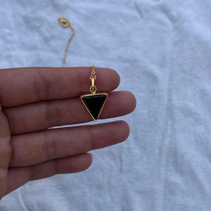 Puede incluir: Un collar dorado con un colgante triangular negro. El colgante cuelga de una fina cadena dorada. La joya se muestra sobre un fondo blanco.