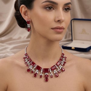 Op de afbeelding: Een sieradenset bestaande uit een ketting en bijpassende oorbellen. De ketting is voorzien van een reeks rechthoekige en peervormige rode stenen, geaccentueerd met kleine heldere steentjes. De oorbellen weerspiegelen dit ontwerp met één steen.