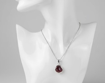 May include: A silver necklace with a pendant featuring a raw, dark red gemstone. The pendant is set in a silver bezel and hangs from a delicate silver chain. The necklace is displayed on a white mannequin bust.