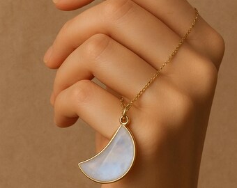 May include: A gold-colored necklace featuring a crescent moon pendant. The pendant has a white and blue iridescent stone set in a gold frame. The necklace is held by a hand against a neutral background.