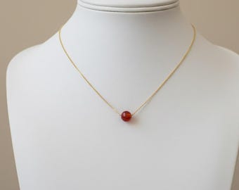 May include: A delicate gold necklace featuring a single, round, reddish-orange gemstone pendant. The necklace is displayed on a white mannequin, showcasing the minimalist design and the vibrant color of the gemstone.