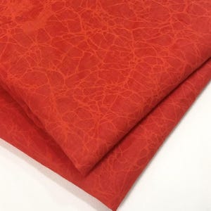 Puede incluir: Una pieza de tela doblada en un tono rojo anaranjado vibrante. El tejido presenta un patrón de grietas distintivo y repetitivo en toda su superficie, que se asemeja a una red de líneas finas interconectadas que crean un efecto visual texturizado.