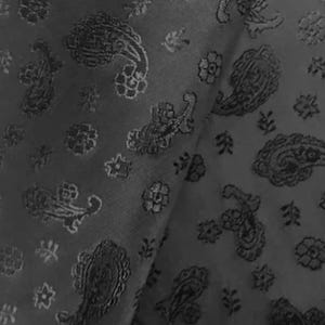 May include: Close-up of a dark grey fabric featuring a paisley pattern. The fabric is folded, showing the texture and design. The paisley motifs are mixed with floral and other decorative elements.