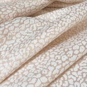 May include: Close-up of folded fabric featuring a cream background with a repeating tan leopard print. The fabric has a textured surface and is soft, with visible folds and shadows.