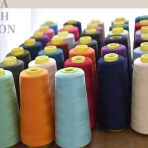 Könnte beinhalten: Eine Sammlung zahlreicher konischer Garnspulen in einer Vielzahl von Unifarben, darunter Hellblau, Orange, Rot, Marineblau und Beige. Jede Spule verfügt über eine leuchtend gelbe Kunststoffkappe und ist in einer gitterartigen Formation angeordnet.