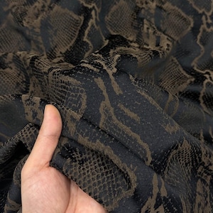 Pode incluir: Um tecido com um padrão de pele de cobra em preto e castanho escuro. O material tem uma superfície texturizada com um design repetitivo de escamas de réptil que cria um efeito visual em camadas sobre a superfície têxtil dobrada e flexível.