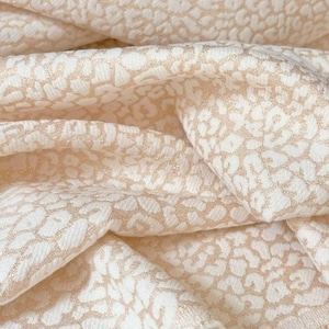 Peut inclure: Un tissu présentant un motif léopard répétitif dans des tons crème et or métallique. Le matériau possède une surface texturée avec des plis et des drapés visibles, mettant en valeur les accents dorés scintillants sur le fond crème mat.