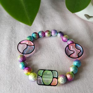 May include: A colorful bracelet featuring iridescent beads in shades of pink, blue, green, and yellow. The bracelet includes two round and one square decorative bead with black outlines and colorful designs. The bracelet is displayed on a light pink surface.